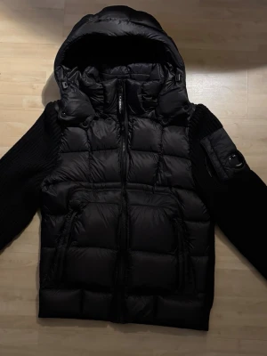 Svart cardigan - Snygg svart västcardigan med stickade ärmar och baksida. Jackan har två stora fickor framtill och en extra ficka med dragkedja på ena ärmen. Puffad kropp i lite glansigt material och ärmar i grovstickad kvalitet ger en cool kontrast. Jackan är ny, har endast testat en gång. Tagsen är såklart kvar men var dum nog och tog av dom. Kom gärna med frågor eller prisförslag!