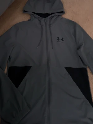  Under Armour - Snygg grå jacka från Under Armour med svart logga på bröstet. Jackan har huva med dragsko, hel dragkedja och svarta detaljer vid fickorna. Materialet är slätt och vattenavvisande, perfekt för blåsiga dagar. Priset kan diskuteras😃