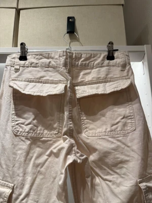 Krämvita cargobyxor med fickor - Säljer ett par beiga cargobyxor med raka ben, low waist och stora fickor både bak och på sidorna. Byxorna har en avslappnad passform och är tillverkade i ett bi bomullstyg. Perfekta för en chill och trendig look. Det finns en lite fläckar från smink men kan gå bort i tvätten. Midjan 36 cm, skrevet till midjan ca 23 cm, inrebenslängd  75cm