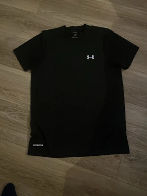 Svart t-shirt från Under Armour - En svart t-shirt från Under Armour med klassisk passform och rund hals. T-shirten har en liten vit logotyp på bröstet och är tillverkad i ett mjukt, funktionsmaterial som andas. Perfekt för träning eller sportaktiviteter.
