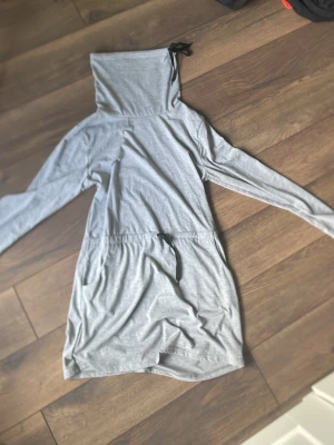 Grå långärmad loungewear-klänning - Mysig grå loungewear-klänning med långa ärmar och hög krage med dragsko. Klänningen har även en dragsko i midjan för justerbar passform. Perfekt för avslappnade dagar hemma.