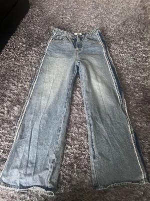 Ljusblå jeans med vida ben - Säljer ett par ljusblå jeans med breda, raka ben och fransiga detaljer längs sidorna. Jeansen har klassisk femficksmodell och är gjorda i denim. Perfekta för en trendig och avslappnad stil.