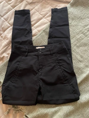 Svarta kostymbyxor från Freequent - Snygga svarta kostymbyxor från Freequent i storlek XXS. Byxorna har raka ben, två knappar i midjan och sidofickor. Perfekta för en clean och stilren look. Materialet känns mjukt och följsamt.