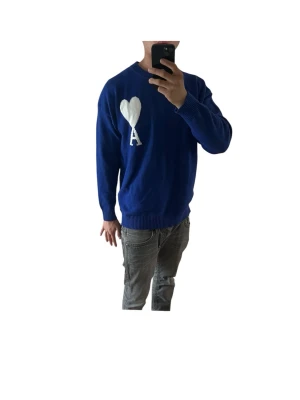Blå AMI Tröja - Snygg blå stickad tröja från AMI i storlek M med vit hjärtdetalj och bokstaven A på bröstet. Tröjan har rund halsringning, ribbade muddar och avslut. Perfekt för dig som vill ha en unik och trendig look. Materialet är  mjukt och bekvämt. Personen i bilden är 180 69kg
