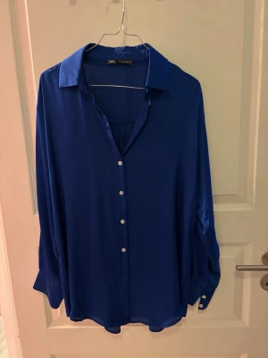 Kornblå satinblus från Zara - En kornblå blus i glansigt satinmaterial från Zara. Blusen har klassisk krage, knäppning framtill med vita knappar och långa ärmar med manschett och knappar. Perfekt för en trendig och stilren look.