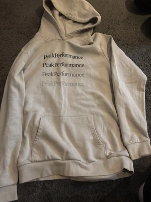 Vit hoodie från Peak Performance - Säljer en vit hoodie från Peak Performance med tryckt logga i flera nyanser på bröstet. Hoodien har en klassisk känguruficka framtill och ribbade muddar vid ärmslut och nederkant. Perfekt för en avslappnad och stilren look.