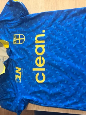 Sverige matchtröja - Säljer en blå sporttröja med mönstrad struktur och tryck i gult. Tröjan har korta ärmar, gul krage och tryck med texten 'clean.' samt loggor på bröstet. Perfekt för träning eller lagidrott.