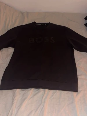 Svart sweatshirt från BOSS - Svart sweatshirt från BOSS med diskret logga i svart på bröstet. Tröjan har rund halsringning och långa ärmar. Tillverkad i mjukt material som känns skönt mot huden. Perfekt för en stilren och avslappnad look.