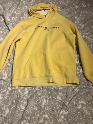 Gul hoodie från Tommy Hilfiger - Snygg gul hoodie från Tommy Hilfiger med tryckt logga och text på bröstet. Klassisk känguruficka framtill och huva med dragsko. Perfekt för en avslappnad och trendig stil. Mjuk bomullskänsla och ribbade muddar vid ärmslut och nederkant.