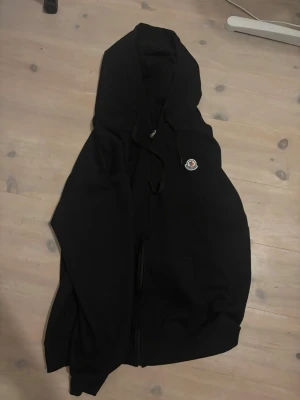 Moncler Hoodie - Säljer en svart hoodie från Moncler med dragkedja framtill och klassisk Moncler-logga på bröstet. Hoodien har justerbar huva med snören och är tillverkad i mjukt bomullsmaterial. Perfekt för en avslappnad och stilren look.