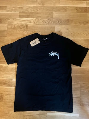 Svart Stüssy t-shirt med logga - Säljer en svart t-shirt från Stüssy med deras ikoniska vita logga tryckt på bröstet. Klassisk passform och rund halsringning. Perfekt till streetwear-stilen och enkel att matcha med allt. Gjord i mjuk bomull för skön känsla. Står M fast passar för S