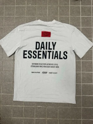 Essentials - Vit t-shirt med stor svart text 'DAILY ESSENTIALS' på ryggen och en liten röd patch upptill. T-shirten har korta ärmar och en klassisk passform. Perfekt för dig som gillar enkel och stilren streetwear.
