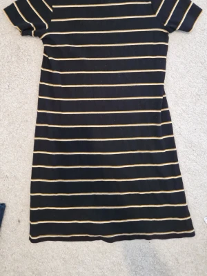 Svart och gulrandig klänning Pull&Bear - Kort svart klänning från Pull&Bear med tunna horisontella ränder i gult och vitt. Klänningen har korta ärmar och rund hals. Materialet känns mjukt och stretchigt, perfekt för en avslappnad stil.