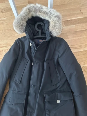 Svart parkas från Woolrich med pälshuva - Snygg svart parkas från Woolrich med stor huva kantad av ljus fuskpäls. Jackan har flera praktiska fickor framtill och stängs med både dragkedja och knappar. Perfekt för kalla dagar och riktigt klassisk streetstil.