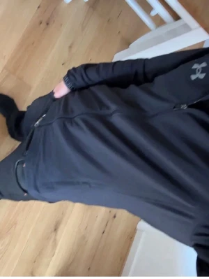 Under Armour windbreaker  - Snygg svart vindjacka från Under Armour med dragkedja framtill, hög krage och logga på bröstet. Jackan har fickor med dragkedja och detaljer med texten 'Under Armour' på ärmsluten. Perfekt för blåsiga dagar och sportiga outfits.