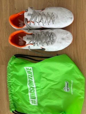 Vita Nike fotbollsskor konstgräs - Säljer ett par vita Nike fotbollsskor med grå snörning och orange insida samt detaljer. Skorna har en låg profil och klassisk Nike Swoosh på sidan. Medföljer en grön gympåse från Unisportlife. Perfekta för dig som vill sticka ut på planen.