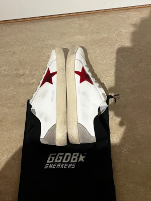 Golden Goose vita sneakers med röd stjärna - Golden Goose sneakers i vitt läder med en ikonisk röd stjärna på sidan och grå detaljer vid hälen. Skorna har beige sula och grå skosnören. Klassisk låg modell med GGDB/SSSTAR-tryck på sidan. Perfekta för en trendig och avslappnad stil.