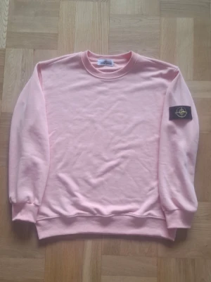 Rosa Stone.island - Ljusrosa hoodie med rund hals och ribbade muddar. Klassisk passform och en svart patch med gult och grönt broderi på vänster ärm. Perfekt för dig som gillar stilrena plagg med en twist.