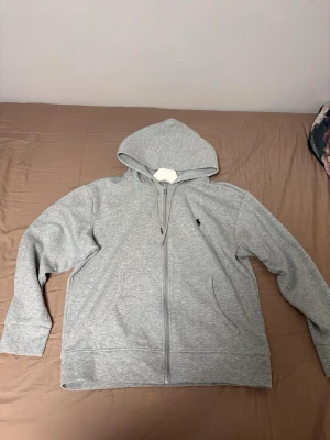 Grå zip hoodie med huva - Säljer en stilren grå hoodie med dragkedja och huva. Hoodien har två fickor framtill och en liten broderad logga på bröstet. Tillverkad i mjukt sweatshirtmaterial, perfekt för en avslappnad look.