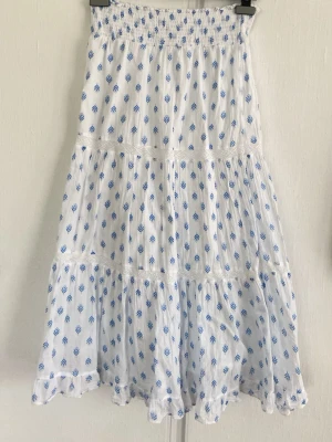 Vit långkjol med blått mönster Gina Tricot - Söt långkjol från Gina Tricot i vitt med blått diskret mönster. Kjolen har smock i midjan, volangdetaljer och är tillverkad i ett lätt och luftigt bomullstyg. Perfekt för varma dagar och ger en fräsch sommarkänsla.