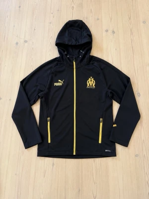 Svart hoodie från Puma med Marseille-logga - Snygg svart hoodie från Puma med gul dragkedja och detaljer. Marseille-logga på bröstet och två fickor med gula dragkedjor. Tillverkad i ett tekniskt material som andas, perfekt för träning eller chill.
