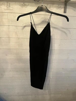 Svart kort klänning med smala axelband - En svart, kort klänning med smala axelband och v-ringning. Klänningen har en enkel och stilren design som passar perfekt för dig som gillar minimalistiskt mode. Materialet är mjukt och följsamt, vilket gör den bekväm att bära.