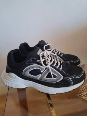 Svarta Dior sneakers med vita detaljer - Snygga svarta sneakers från Dior med vita och svarta detaljer samt vita skosnören. Skorna har en chunky vit sula och stora CD-loggor på sidan. Materialet är en mix av mesh och syntet, vilket ger en sportig och modern look.