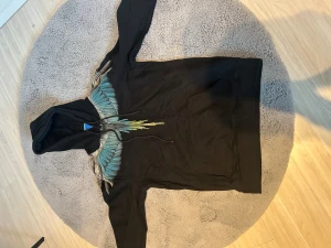 Svart hoodie Marcelo Burlon med vingar - Svart hoodie från Marcelo Burlon County of Milan med ikoniskt vingtryck i blått, grått och gult över axlar och huva. Tröjan har huva med snörning och stor magficka framtill. Perfekt för dig som vill sticka ut med en unik streetwear-look.
