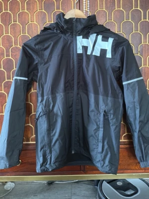 Svart regnjacka från Helly Hansen - Snygg svart regnjacka från Helly Hansen med stor HH-logga på bröstet. Jackan har huva, hel dragkedja framtill, två sidofickor med dragkedja och reflexdetaljer på ärmarna. Tillverkad i vattenavvisande material, perfekt för regniga dagar.