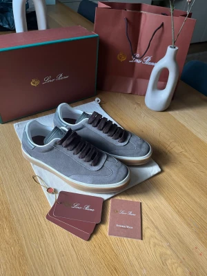 Grå mockasneakers från Loro Piana - Snygga sneakers i grå mocka från Loro Piana med bruna skosnören och vit sula med beige kant. Klassisk låg modell med stilren design och exklusiv känsla. Perfekta för dig som gillar premium och tidlös stil.