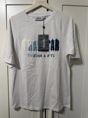 Vit t-shirt från Trapstar  - Vit t-shirt med broderad text i olika blå nyanser på bröstet och trycket 'IT'S A SECRET'.  Den passar som            den passar som S/M