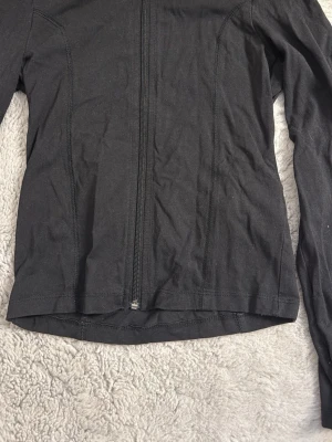 Svart träningsjacka med tumhål - Säljer en svart träningsjacka med hel dragkedja framtill och långa ärmar med tumhål. Jackan är figursydd och tillverkad i ett stretchigt material som passar perfekt för träning eller löpning.