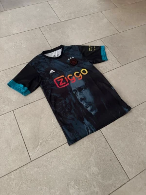 Ajax x Bob Marley svart matchtröja - Säljer en svart Ajax matchtröja från Adidas med Bob Marley-motiv på framsidan. Tröjan har färgglad Ziggo-logga, klubbmärke med tre stjärnor och turkosa detaljer på ärmarna. Perfekt för dig som älskar fotboll och musik!