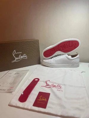 Vita sneakers med nitar från Louboutin👟 - Snygga vita sneakers från Christian Louboutin med ikonisk röd sula och coola nitar framtill. Skorna har en låg profil och är tillverkade i slätt läder. Perfekta för dig som vill sticka ut med exklusiv design och detaljer. Har slitit dem så som man vill ha  de.