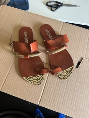 Laura Clement Sandaler - Säljer ett par bruna sandaler från Laura Clement med breda remmar i läderlook och snygga guldfärgade metallringar. Sandalerna har öppen tå och ett unikt mönster i svart och gult framtill på sulan. Perfekta för dig som vill ha något trendigt och annorlunda.