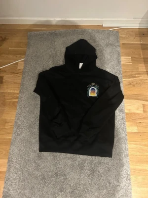 Svart hoodie med Casablanca-tryck - Säljer en svart hoodie med huva och ett färgglatt tryck på bröstet där det står 'Avenida Casablanca' och ett motiv av en byggnad i fönster. Hoodien har ribbade muddar och är i mjukt material, perfekt för en chill och stilren look.