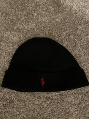 Svart mössa från Polo Ralph Lauren - Snygg svart mössa från Polo Ralph Lauren med klassisk röd logga broderad framtill. Mössan har vikt kant och är ribbstickad nedtill för extra stil och passform. Perfekt accessoar för kyliga dagar.
