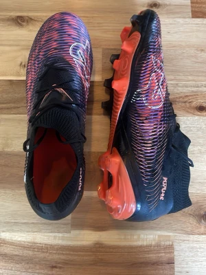 Svarta och röda fotbollsskor Puma - Säljer ett par snygga fotbollsskor från Puma i svart och rött med coolt mönster och orange detaljer. Skorna har snörning, låg profil och greppvänlig sula med dobbar. Perfekta för dig som vill sticka ut på planen! Som nya använda en två gånger inga tecken på användning för endast 199 