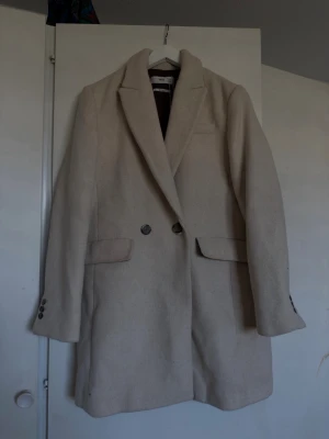 Beige lång kappa med knappar - Stilren beige kappa i längre modell med dubbelknäppning framtill och klassisk krage. Kappan har två fickor med lock och dekorativa knappar vid ärmsluten. Perfekt för dig som gillar en clean och tidlös look.
