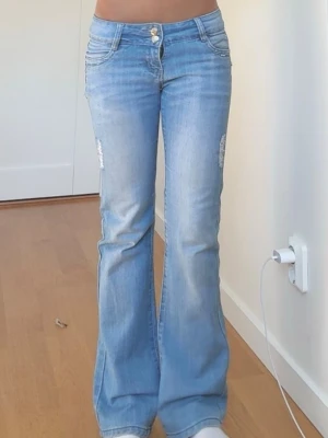 Ljusblå bootcut jeans med slitningar - Säljer ett par ljusblå bootcut jeans med snygga slitningar på låren. Jeansen har hög midja, klassiska fem fickor och stängs med dragkedja och två knappar. Perfekta för dig som gillar en retro vibe och vill ha ett par jeans med lite extra attityd.