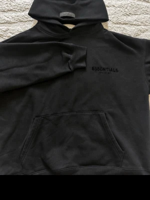 Svart Essentials Fear of God hoodie - Säljer en svart hoodie från Essentials Fear of God med diskret logga på bröstet och stor text på ryggen. Hoodien har en klassisk känguruficka och huva. Perfekt för dig som gillar streetwear och vill ha en stilren look.