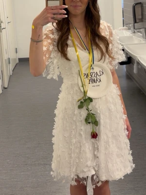 Vit kort spetsklänning med blommor - Superfin vit kort klänning med v-ringning och genomskinliga spetsärmar. Klänningen har 3D-blommor över hela tyget och volangig kjol. Perfekt för student eller sommarfest! Använd endast 1 gång.