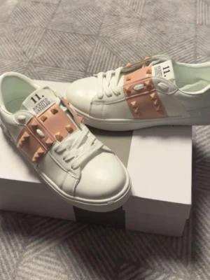 Vita sneakers med rosa nitar - Vita sneakers från Valentino med breda ljusrosa band och pyramidformade nitar över vristen. Skorna har en låg profil, vita snören och en chunky sula med rutmönster undertill. Tillverkade i slätt läder för en clean och modern look.