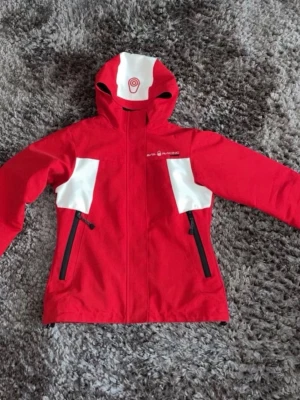 Röd vindjacka från Sail Racing - Snygg röd vindjacka från Sail Racing med vita detaljer på bröstet och huvan. Jackan har två svarta dragkedjeförsedda fickor framtill och justerbar huva. Perfekt för blåsiga dagar och sportiga outfits. Materialet är slitstarkt och vattenavvisande.