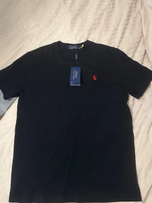 2-pack t-shirts från Polo Ralph Lauren - Säljer två klassiska t-shirts från Polo Ralph Lauren. Ena är ljusgrå med svart logga på bröstet, den andra är svart med röd logga. Båda har rund hals, korta ärmar och är gjorda i mjuk bomull för maximal komfort. 