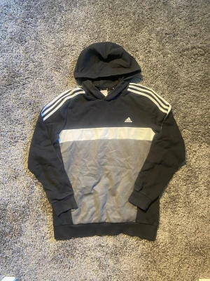 Svart, vit och grå hoodie från Adidas - Snygg hoodie från Adidas i svart och grått med klassiska vita ränder på axlarna och en vit panel över bröstet. Tröjan har huva och ribbade muddar vid ärmslut och nederkant. Perfekt för en sportig och avslappnad stil.