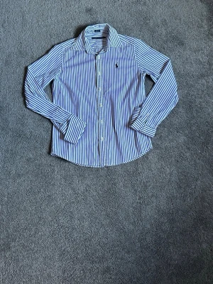Blåvit randig skjorta från Ralph Lauren - Klassisk blåvit randig skjorta från Polo Ralph Lauren med broderad logga på bröstet. Skjortan har lång ärm, knappar framtill och är tillverkad i mjuk bomull. Perfekt för en stilren och tidlös look.