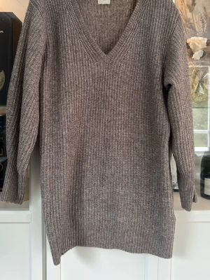 Grå stickad klänning med v-ringning - Mysig grå stickad klänning med djup v-ringning och lång ärm. Klänningen har en rak passform och ribbstickad struktur som ger en avslappnad look. Perfekt för lager på lager och enkel att matcha med olika outfits.
