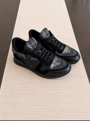 Valentino Garavani Rockrunners - Snygga svarta sneakers från Valentino med grått kamouflagemönster och nitar på sulan. Skorna har detaljer i mocka, läder och textil samt bronsfärgade partier vid hälen. Perfekta för dig som vill ha en unik och trendig look.