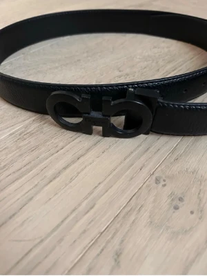 Svart skärp med mattsvart spänne - Snyggt svart skärp i skinn med ett stilrent mattsvart metallspänne i en unik design. Perfekt accessoar för att lyfta din outfit med en modern touch. Skärpet har en slät yta och klassisk form. Ferregamo